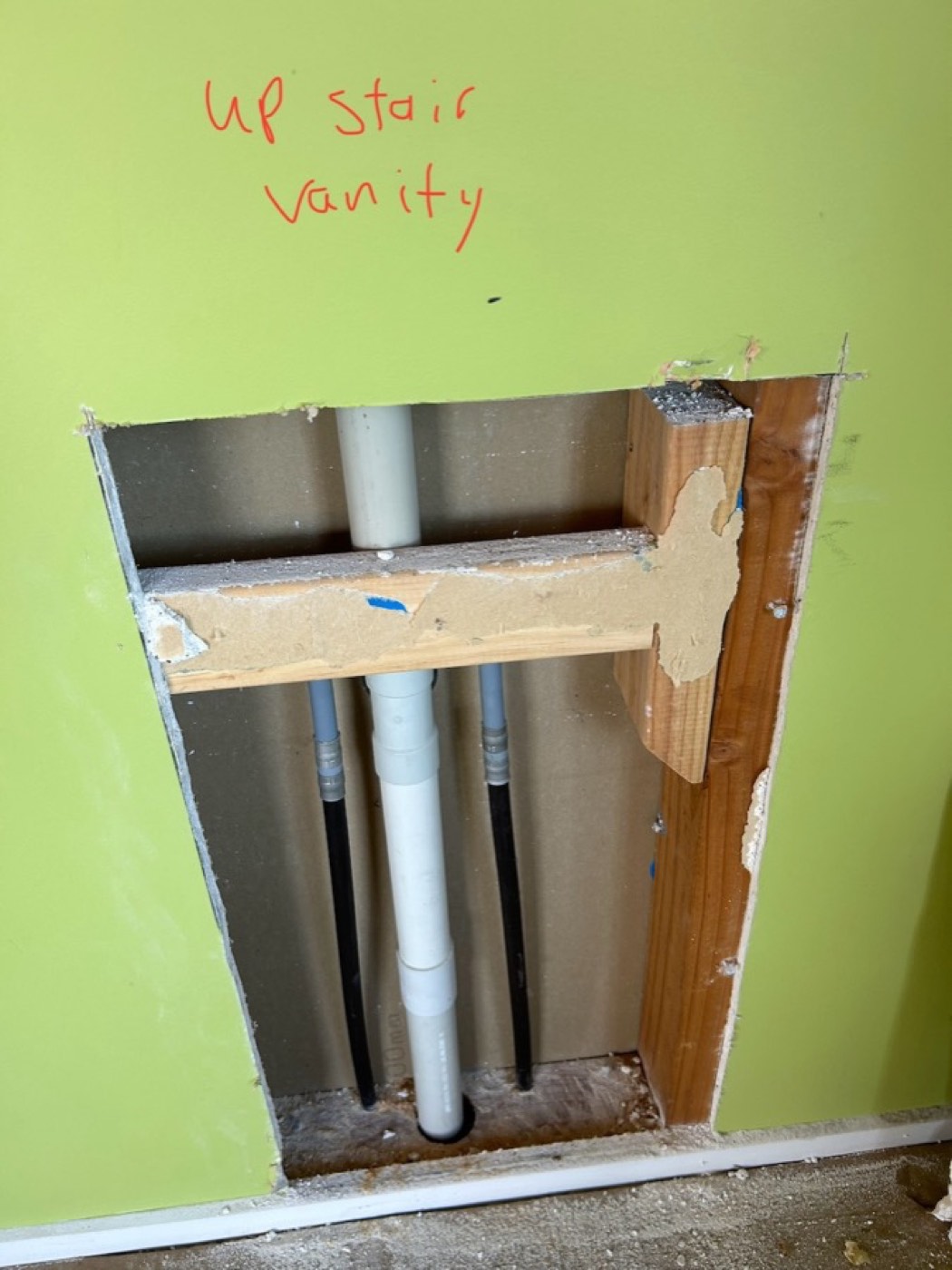 A wall opened up behind an upstairs vanity during a house re-pipe, with old black pipe and the new pipework in the cavity, and the location written on the wall in marker